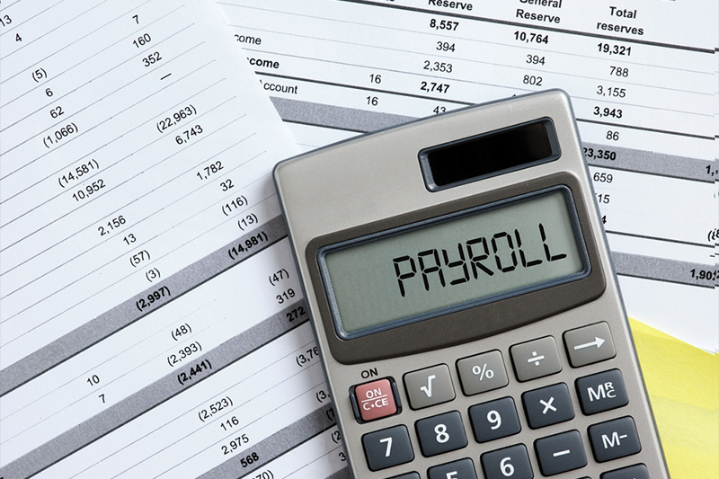 A calculator with the word 'PAYROLL' on the screen, sitting on top of financial spreadsheets.